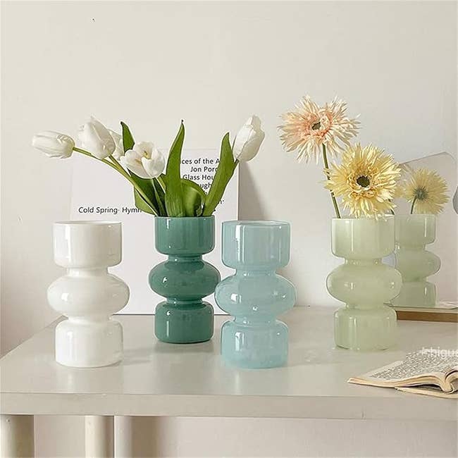 Four glass vases in white, light blue, light green, and dark green holding flowers