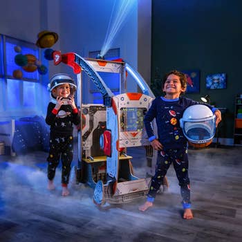 Two child models with space helmets and pajamas standing in front of wooden space ship set