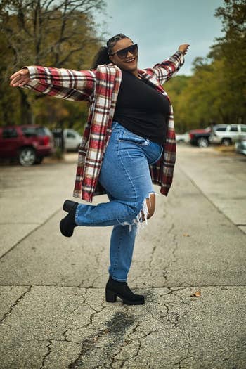 Person joyfully posing outdoors, wearing a plaid coat, achromatic top, ripped jeans, and boots, suggesting a casual, stylish look for autumn