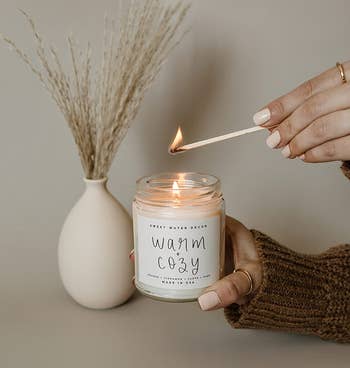 hands lighting the 'warm + cozy' candle