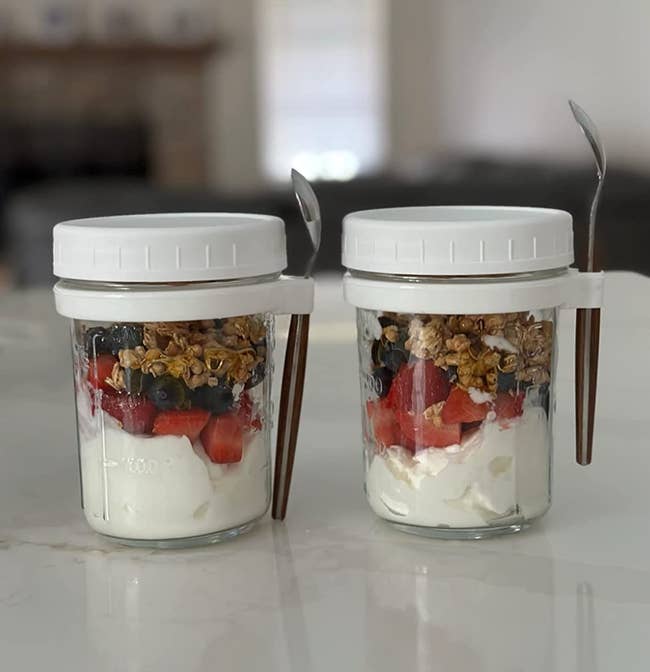Two small cylinder jars full of yogurt and granola with twist off white caps and a tiny spoons attached to them 