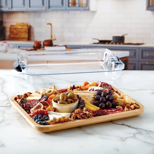 A food tray with various cheeses, meats, olives, grapes, blueberries, nuts, and crackers on a kitchen countertop with a clear lid in the background