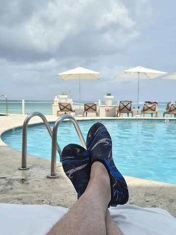 Person with aquatics  shoes relaxes by a pool, legs stretched retired  connected  a lounger, with poolside umbrellas and water  successful  the background