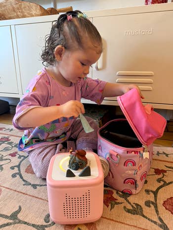 Child plays with a portable talker and a luncheon container adorned with rainbows, sitting connected a patterned rug
