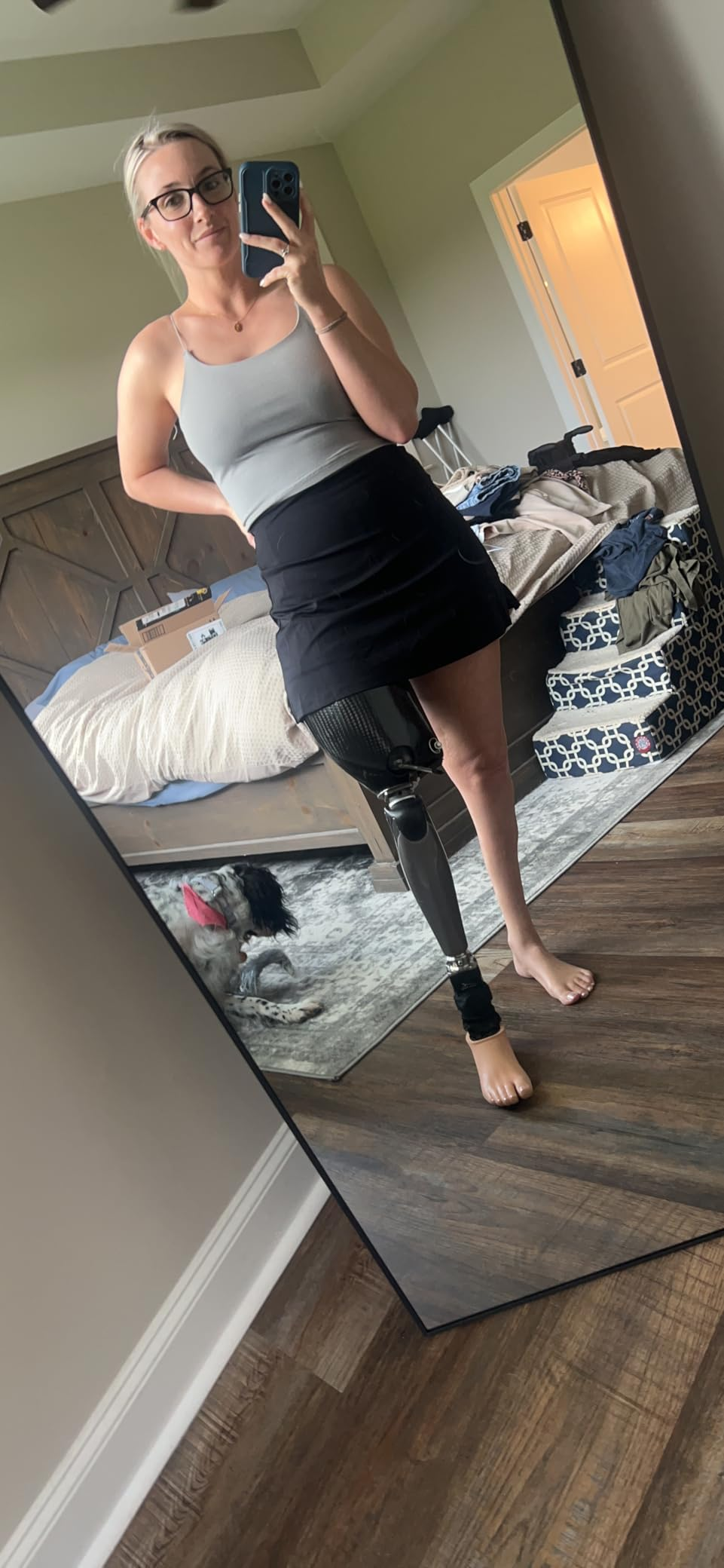 A woman with a prosthetic leg takes a mirror selfie wearing a tank top and skirt, standing in a bedroom