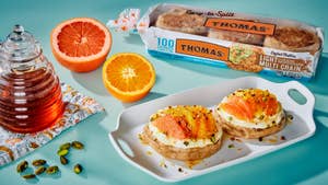 A blue tabletop with two English muffins topped with cream and sliced citrus fruits on a white platter, two citrus halves, a glass jar of honey on a tablecloth, and a package of English muffins.