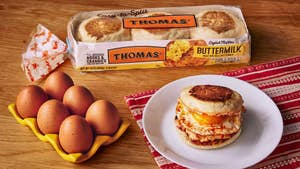 A tabletop with six whole eggs in a yellow carton, an egg muffin sandwich on a white plate and a red-and-white placemat, and a package of English muffins. 