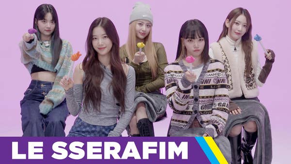 Chaewon, Sakura, Yunjin, Kazuha, and Eunchae of Le Sserafim point at the camera with colorful presentation sticks that they use throughout the video to point out their picks for their answers to BuzzFeed's who's who questions.