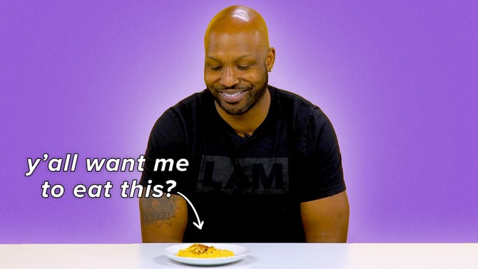 Phil sits smiling in a black tee shirt looking at a plate of yellow shrimp and grits. The text reads, 