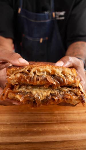 Two hands hold a grille cheese sandwich with caramelized onions, over a wooden table.