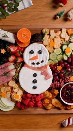 snowman cheeseboard with snowman made out of brie, proscuitto scarf, berries as eyes and buttons, and a carrot nose, surrounded by other charcuterie board staples