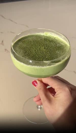 Hand holding a coupe glass filled with a frothy green drink