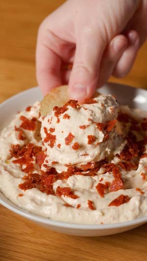 Closeup of a potato chip being dipped in a creamy, thick dip with chopped pepperoni on top.