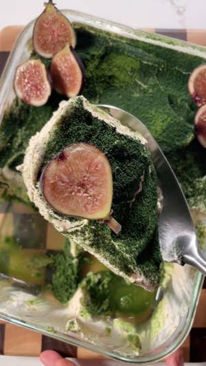 Closeup of a metal spoon scooping white cream, topped with green matcha powder and a slice of fresh fig, from a baking dish.
