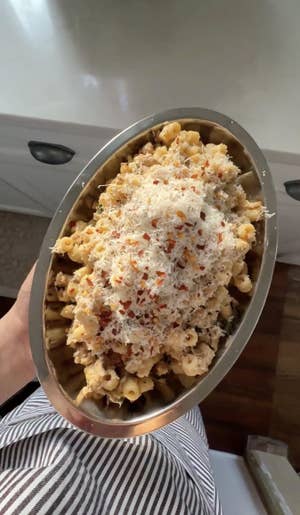 Hand holding up a metal oval-shaped platter filled with cheesy pasta tubular noodles, topped with grated Parmesan cheese and red pepper flakes