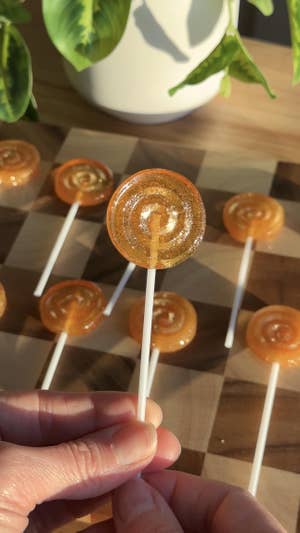 Two hands holding up a round golden lollipop on a white lollipop stick, with a backdrop of a brown checkered cutting board topped with more lollipops.