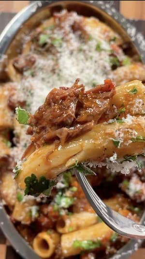 Closeup of beef ragu with rigatoni noodle on a fork, with a baking dish of ragu rigatoni in the background