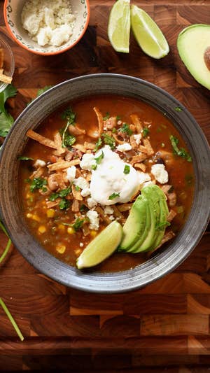 Metal round bowl filled with red broth, corn, beans, shredded chicken, sliced avocado, crumbled white cheese, sour cream, and chopped green herbs.