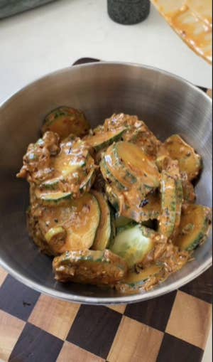 Round metal bowl filled with peanut sauce-coated cucumber slices, sitting on a brown checkered cutting board