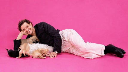 Pedro Pascal lays in front of pink background with some puppies.