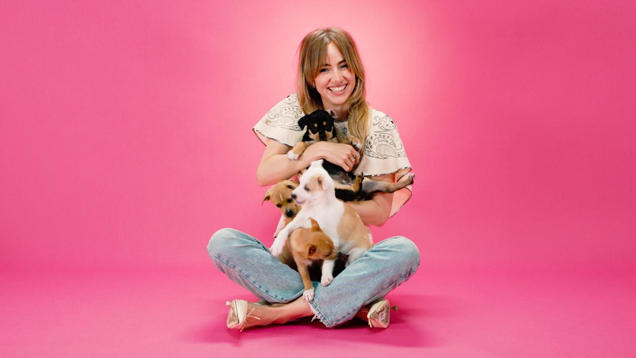 Suki Waterhouse sits in front of a pink background holding puppies.